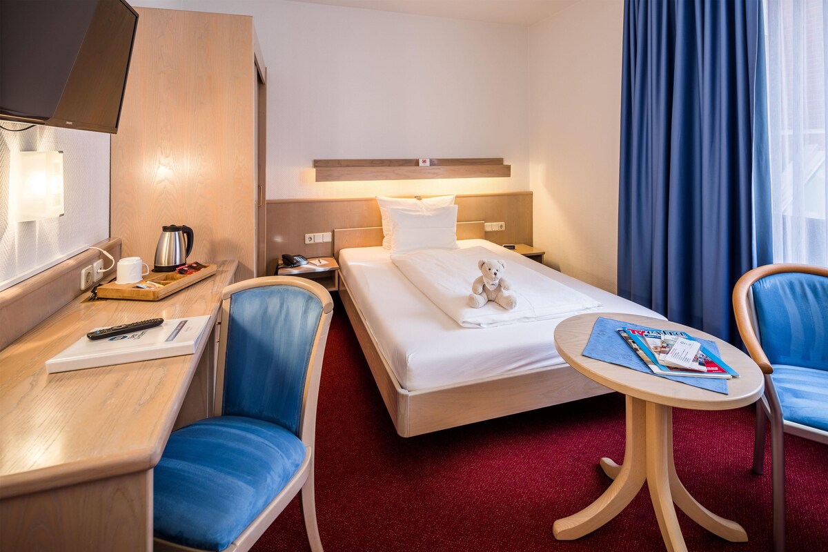 A hotel room with a bed, a desk with a chair, a small table with a chair, a teddy bear on the bed, and a magazine on the table.