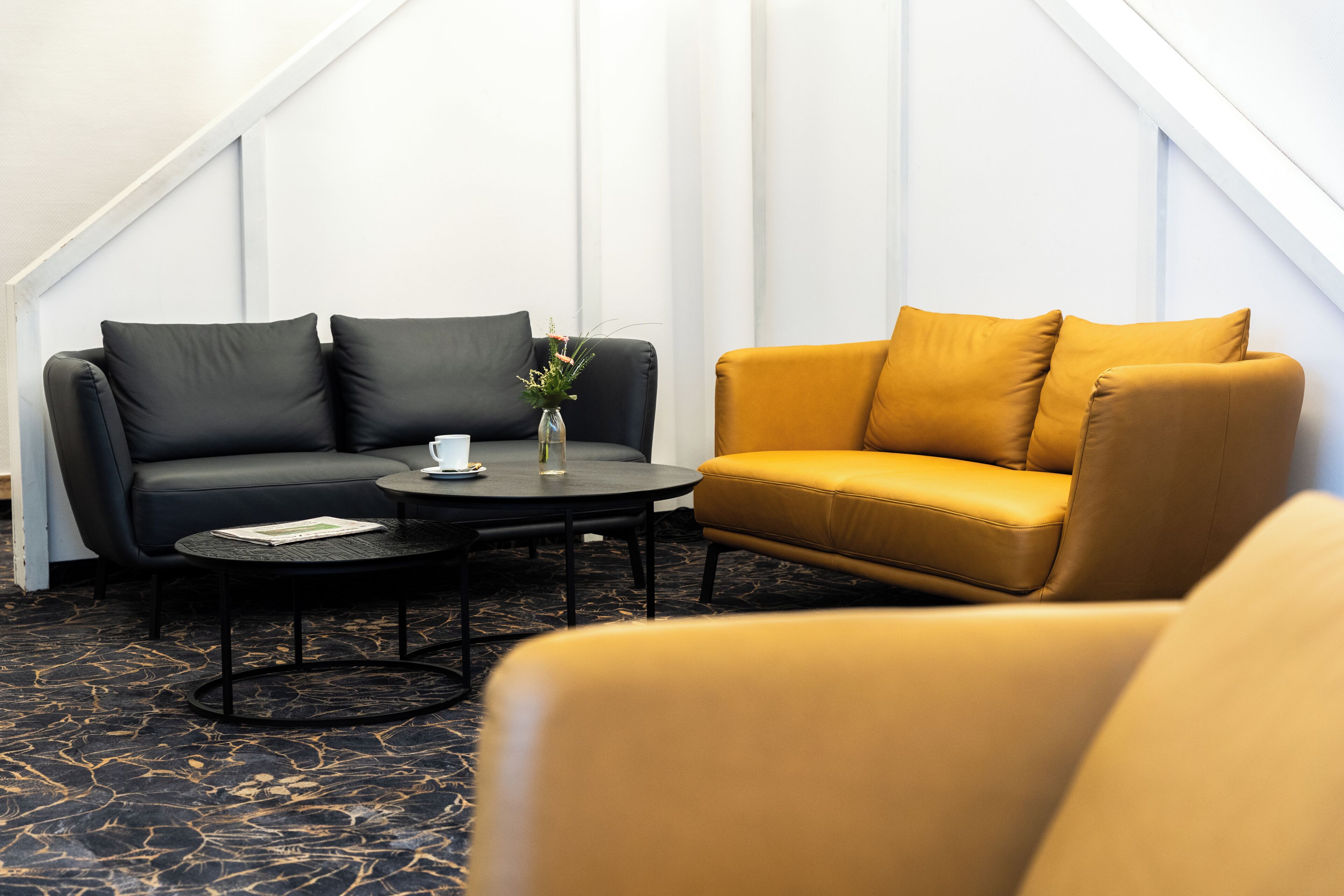 Une pièce avec un canapé noir, un canapé jaune, une table basse ronde et un vase rempli de fleurs.