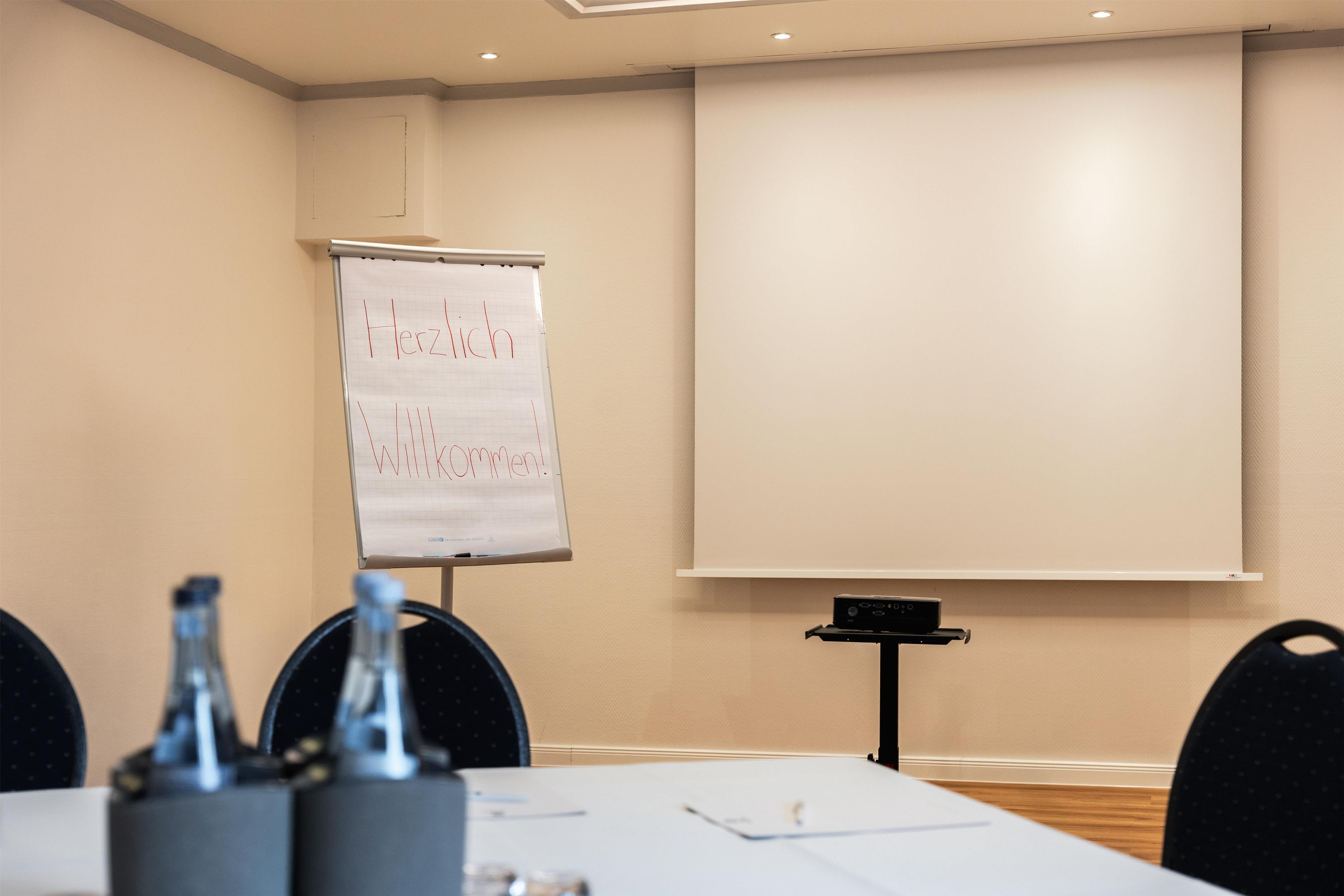 Ein Konferenzraum mit einer Whiteboard, einer Projektionsleinwand und einem Tisch mit Stühlen.