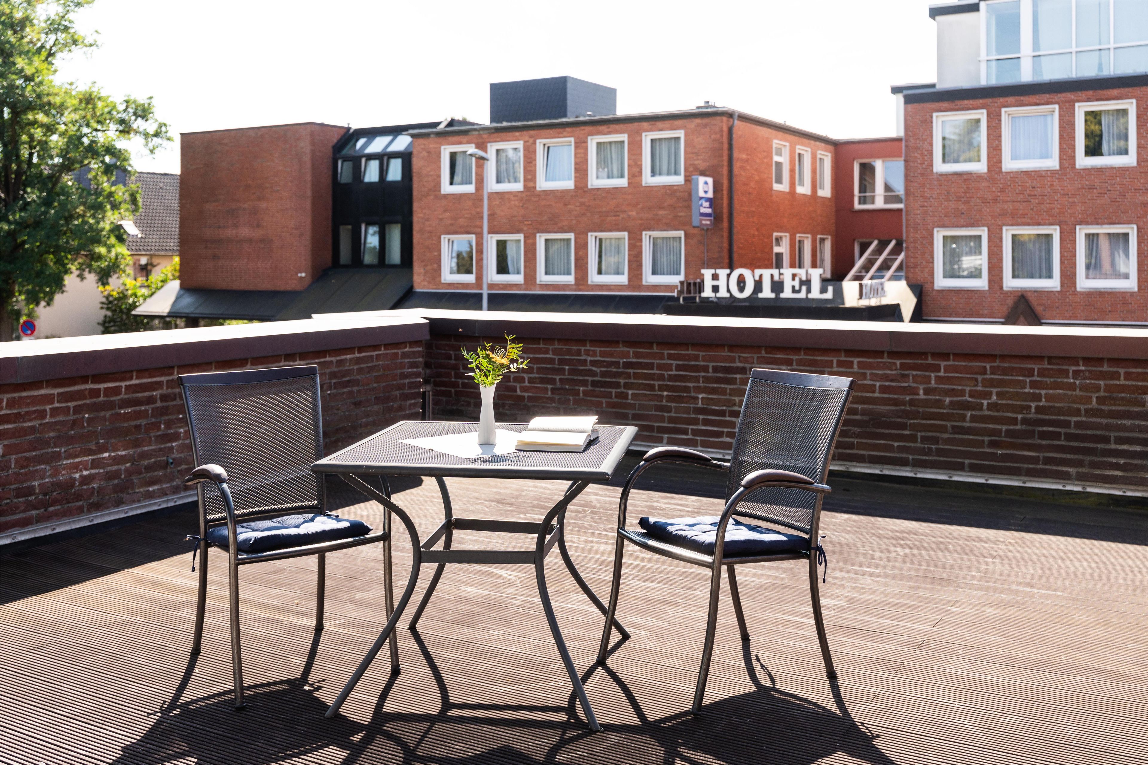 En utomhusplats med sittmöjligheter, bestående av två metallstolar och ett bord, en krukväxt och en skylt som visar att det finns ett hotell här.