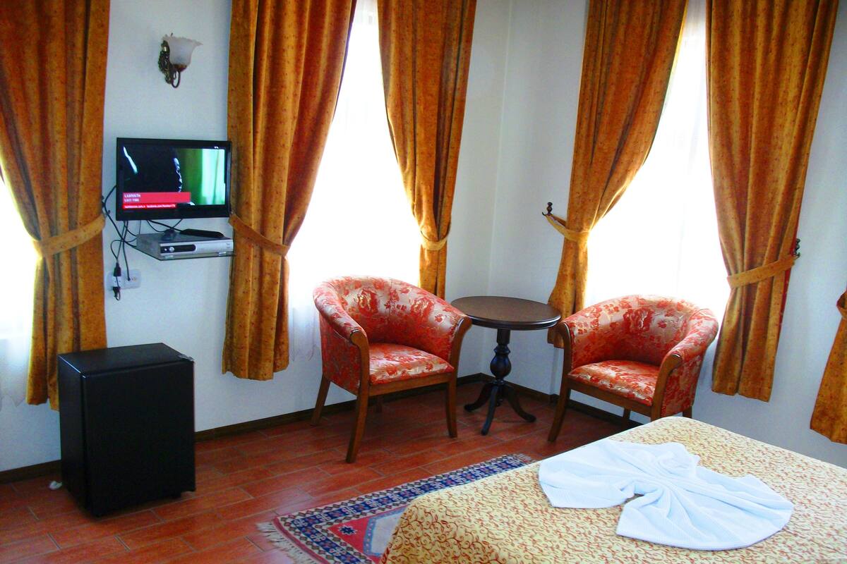 A hotel room with a bed, two armchairs, a small table, a TV, and a speaker.