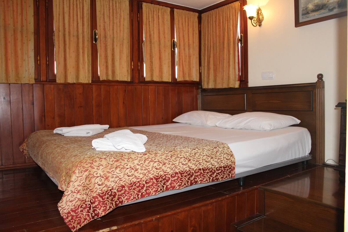 A neatly made bed with a patterned bedspread and white pillows, wooden paneling on the walls, and a wall-mounted light fixture.