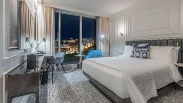 A hotel room with a large bed, a desk, a chair, and a view of the cityscape at night.