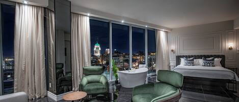 A modern hotel room with a large bed, green armchairs, a round table, a bathtub, and a city view.