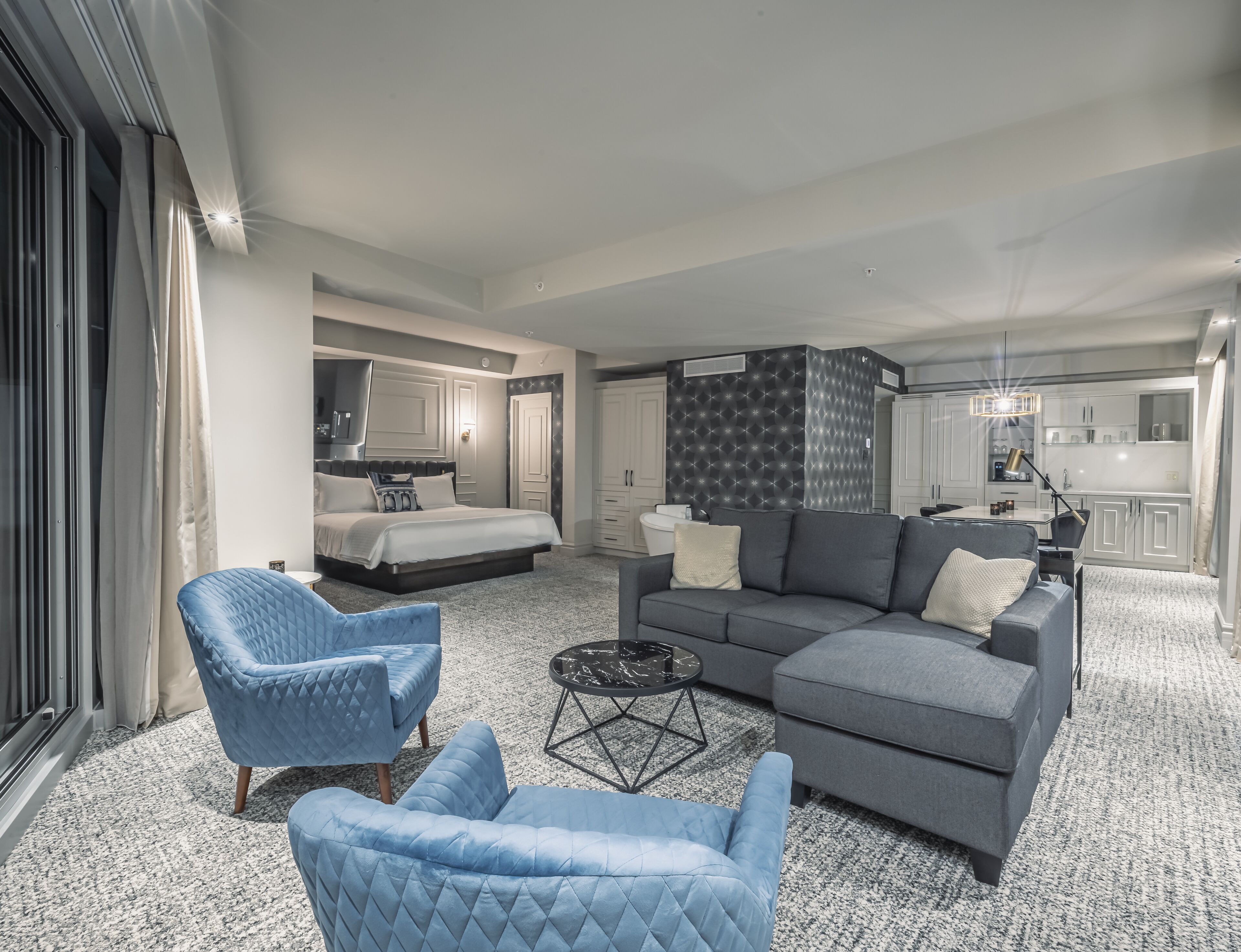 Una habitación de hotel moderna con una cama grande, un sofá gris, un sillón azul y un comedor.