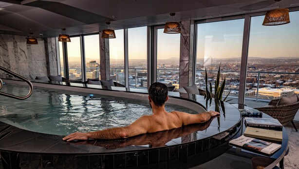 A man relaxing in a hot tub with a city view, a lounge area with seating, and a table with books and a plant. - Le Capitole Hôtel (Québec City)