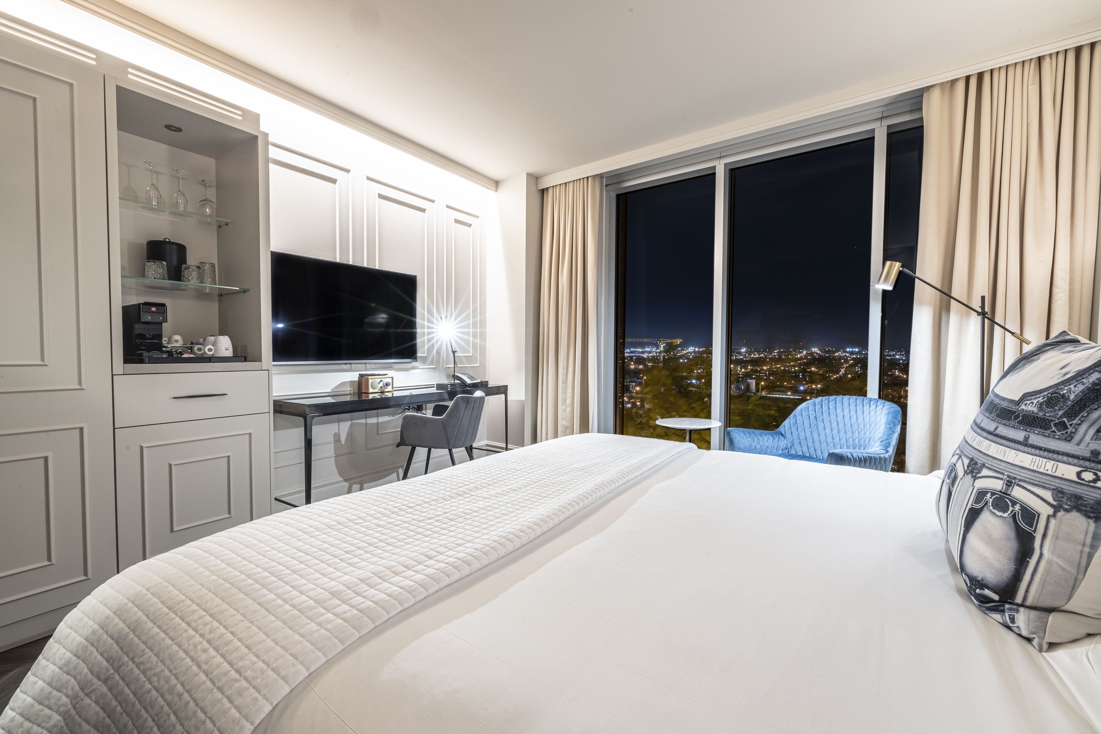 Una camera d'albergo con un letto grande, una scrivania con televisore a schermo piatto, una sedia e vista sulla città di notte.