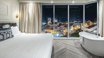 Et moderne hotelværelse med en stor seng, et badekar og udsigt over byen gennem gulv til loft-vinduer.
