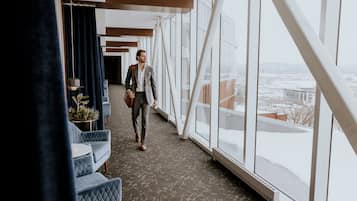 Un pasillo moderno de hotel con amplios ventanales, vistas a la ciudad y una zona de estar con sillones tapizados en azul.