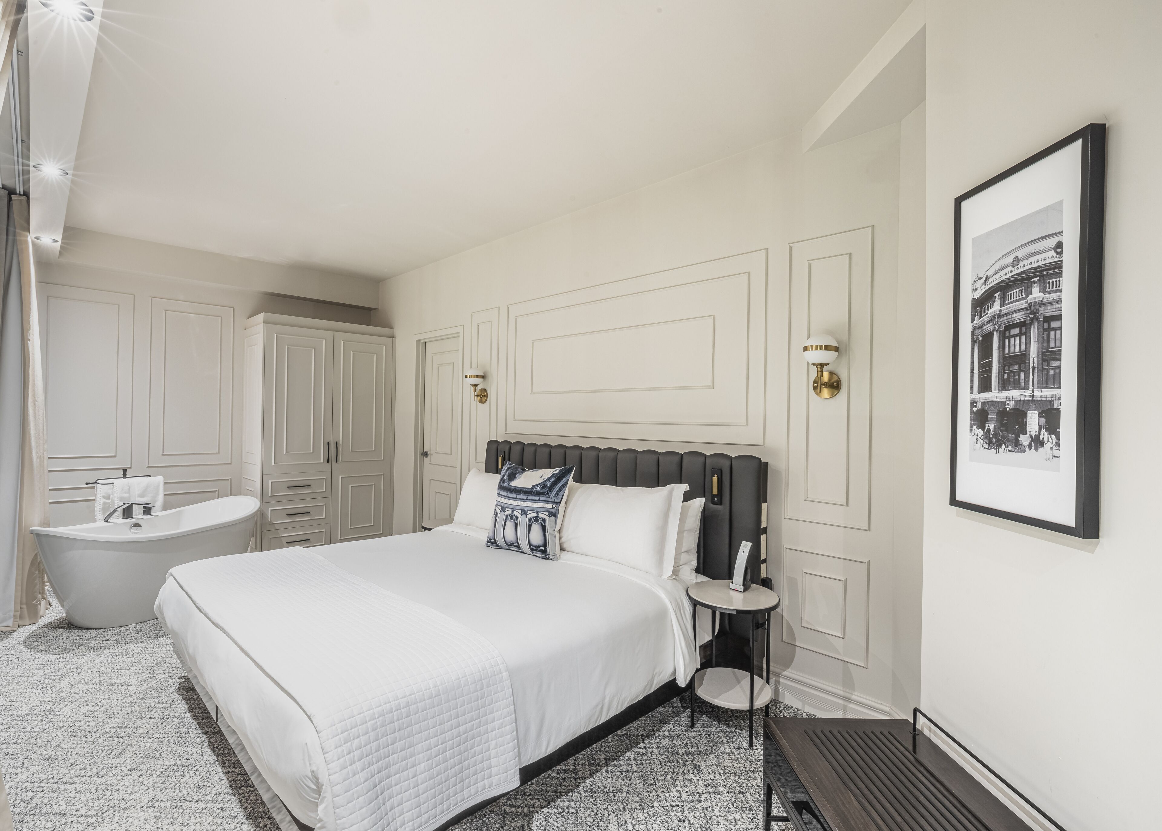 Une chambre à coucher avec un grand lit, une table de chevet, une commode, une lampe fixée au mur, un tableau encadré, une baignoire et une armoire.