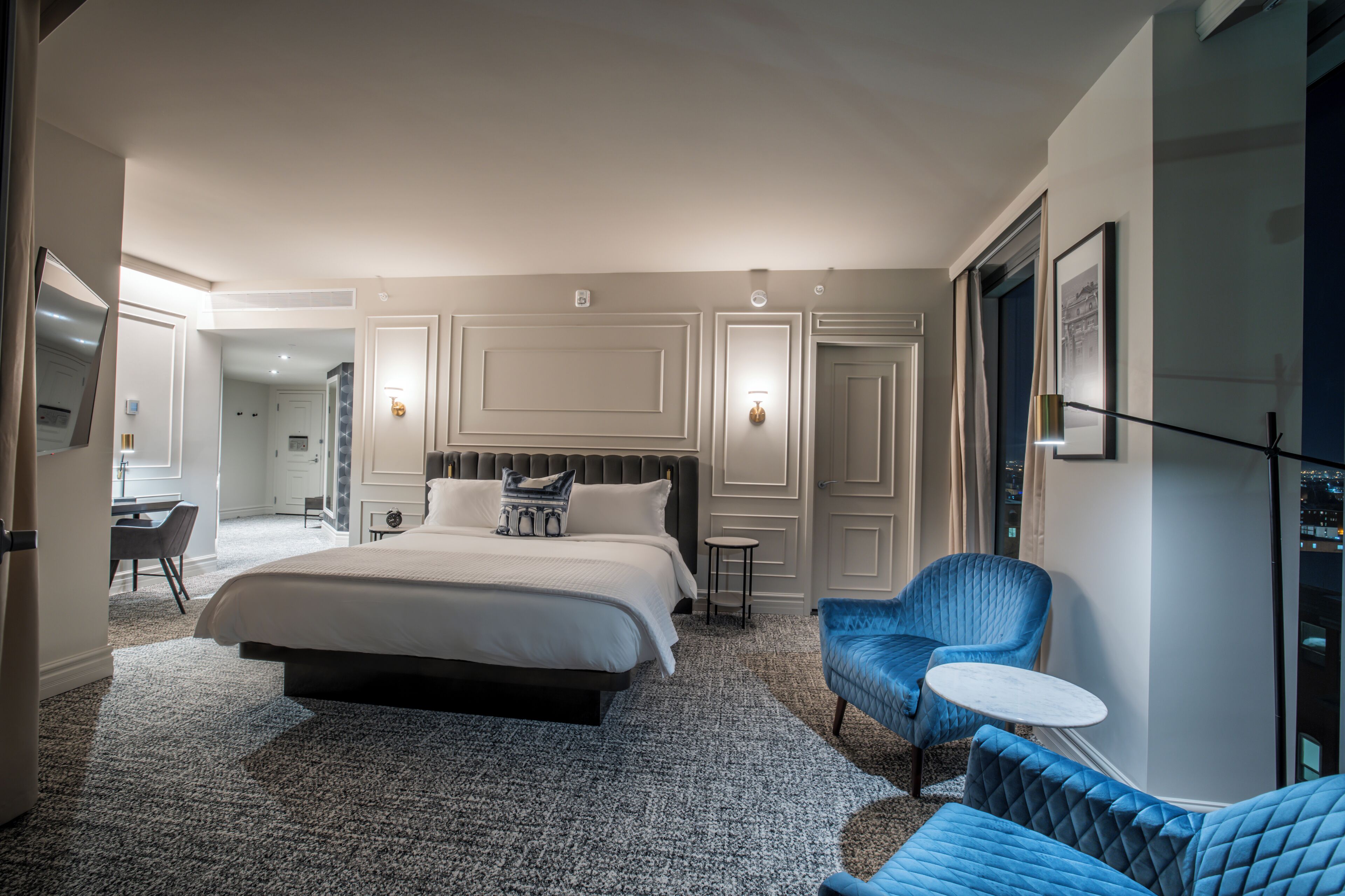 Una habitación de hotel moderna con una cama grande, una zona de estar con una silla azul y una mesita, y vistas a la ciudad desde la ventana.
