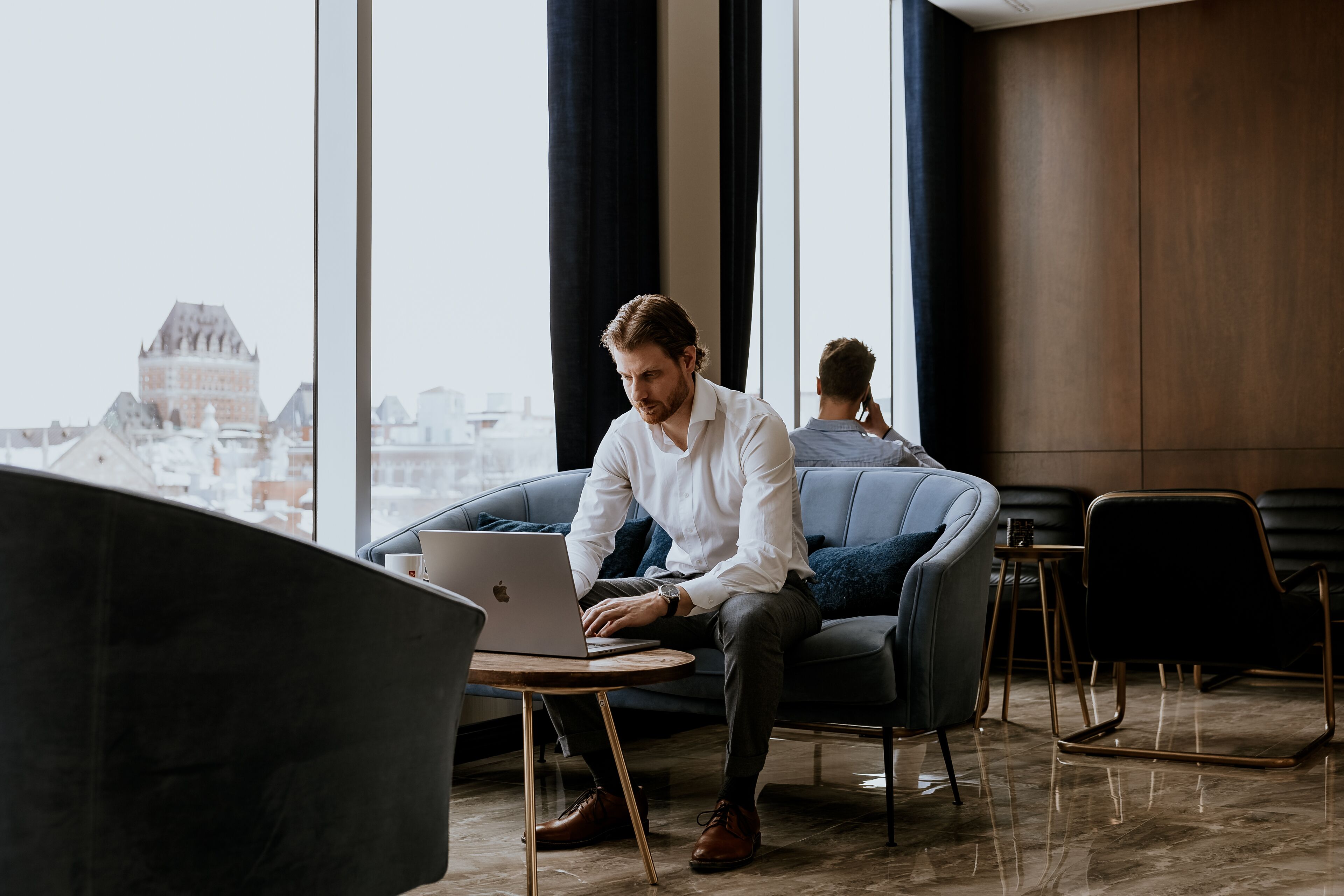 Ein Mann arbeitet an einem Laptop in einem modernen Büro mit großen Fenstern, Blick auf ein historisches Gebäude und zeitgenössischen Möbeln.