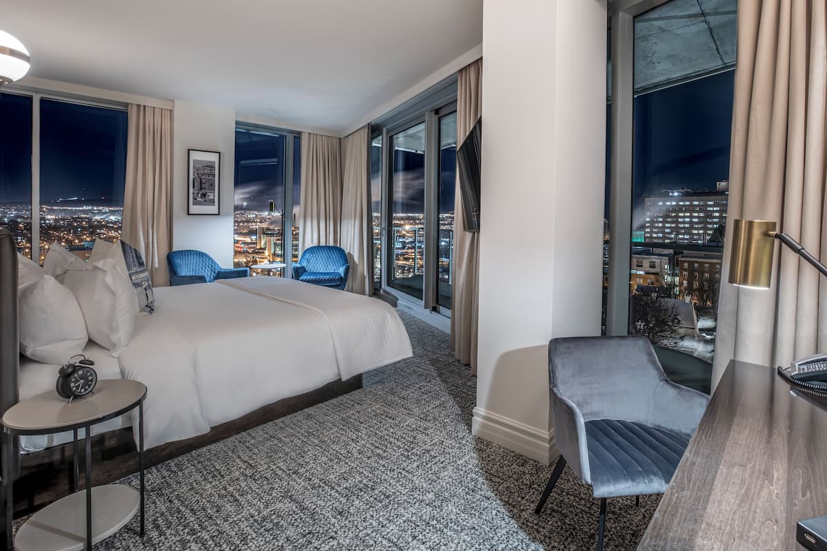 Hotellihuone, jossa on suuri sänky, kaksi sinistä nojatuolia, työpöytä lampun kanssa ja näkymä kaupunkiin yöllä.