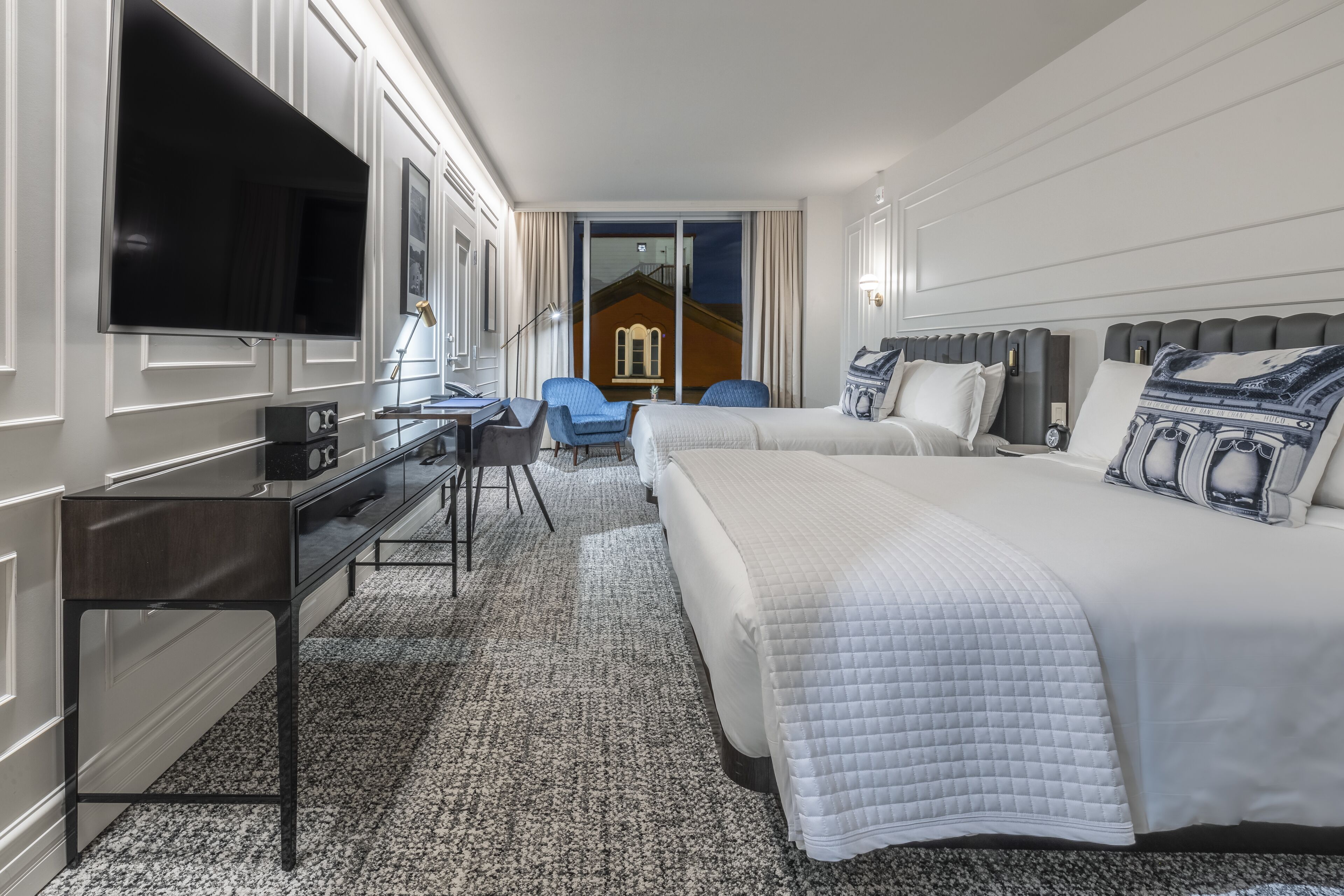 A hotel room with a large bed, a desk with a computer, a TV, and a view of a building through the window.