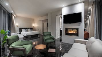 A modern hotel room with a fireplace, a flat-screen TV, a bed, a dining area, and a seating area with green chairs and a white sofa.