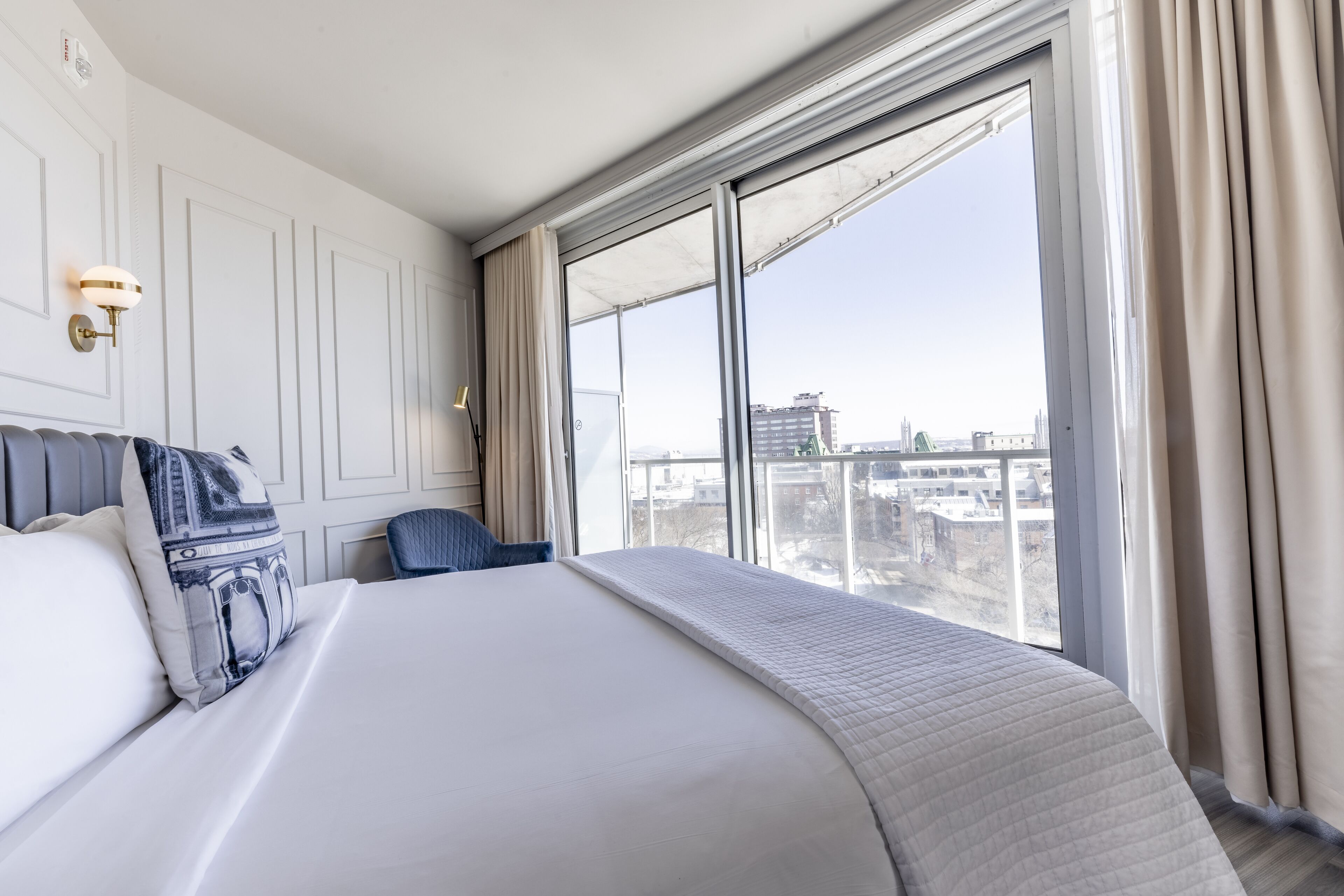 A hotel room with a large bed, a chair, a nightstand, a lamp, and a window with a city view.