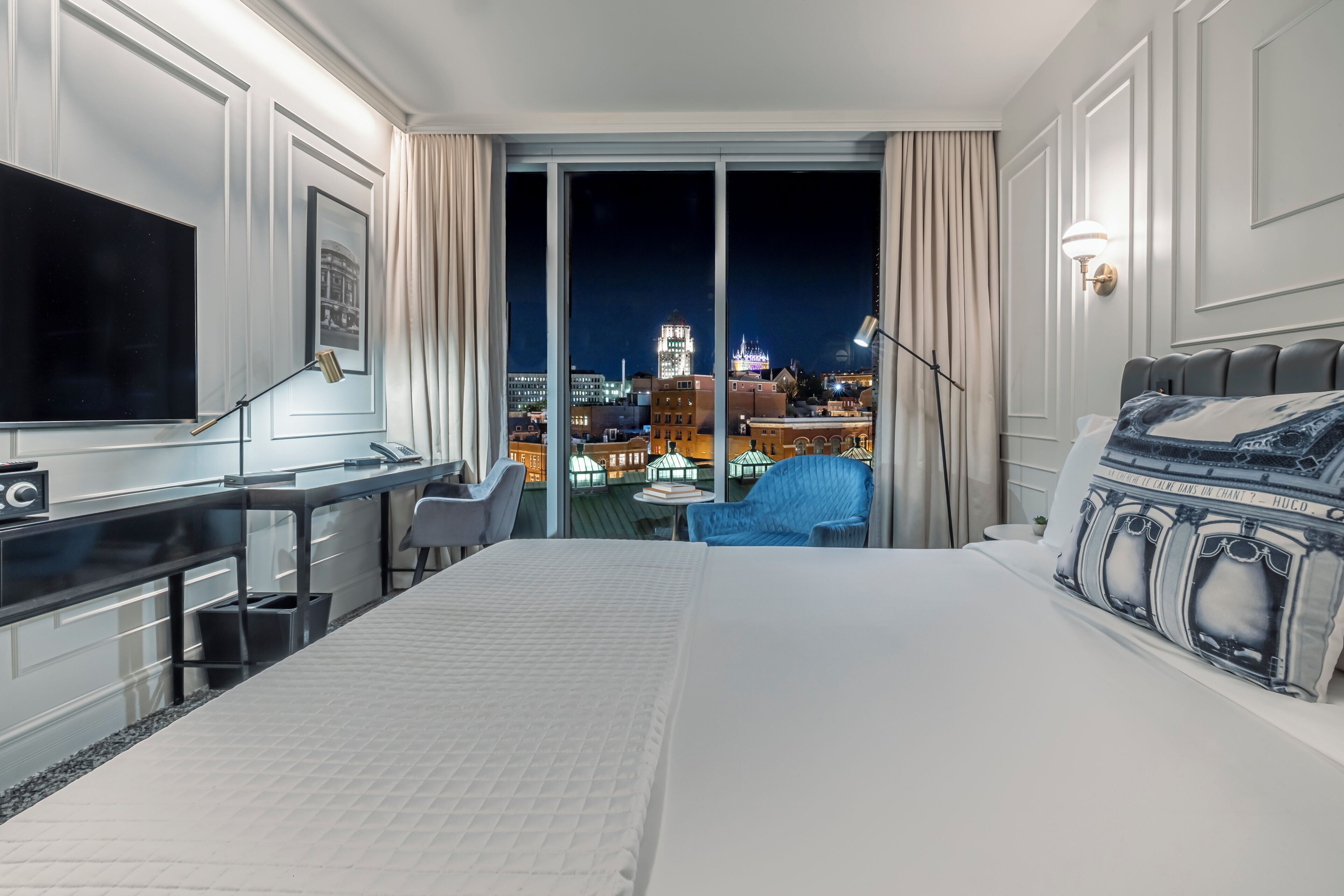 A hotel room with a large bed, a desk, a chair, a TV, and a view of a cityscape at night.