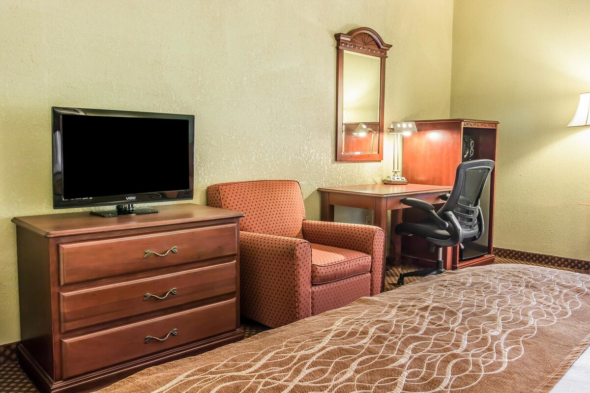A hotel room with a TV, a wooden dresser, a patterned bedspread, a polka-dotted armchair, a desk with a lamp, and a mirror.