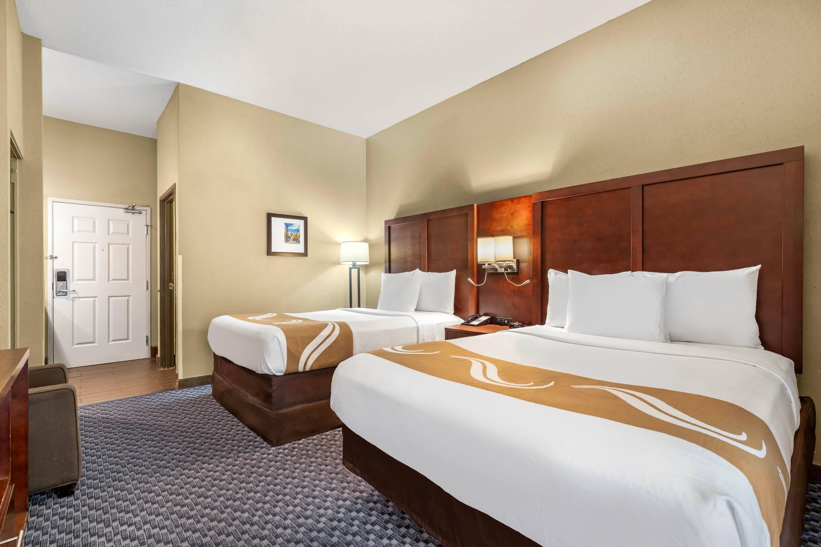 A hotel room with two beds, a desk, a chair, a door, and a framed picture on the wall.