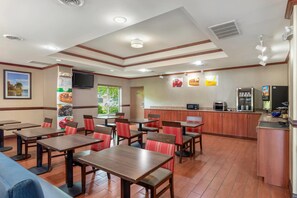 Un restaurante con mesas y sillas de madera, una zona de cocina y un televisor montado en la pared.