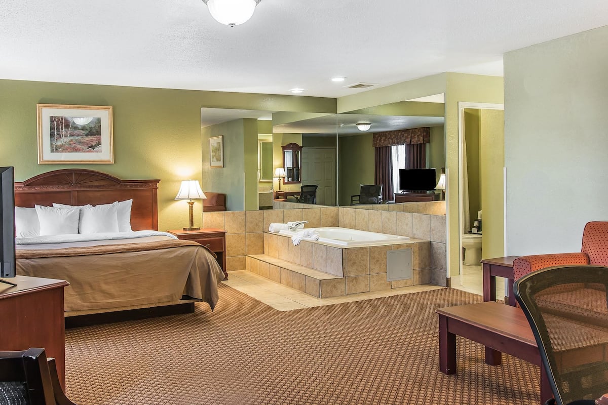 A hotel room with a bed, a sitting area, a large mirror, a bathtub, and a television.