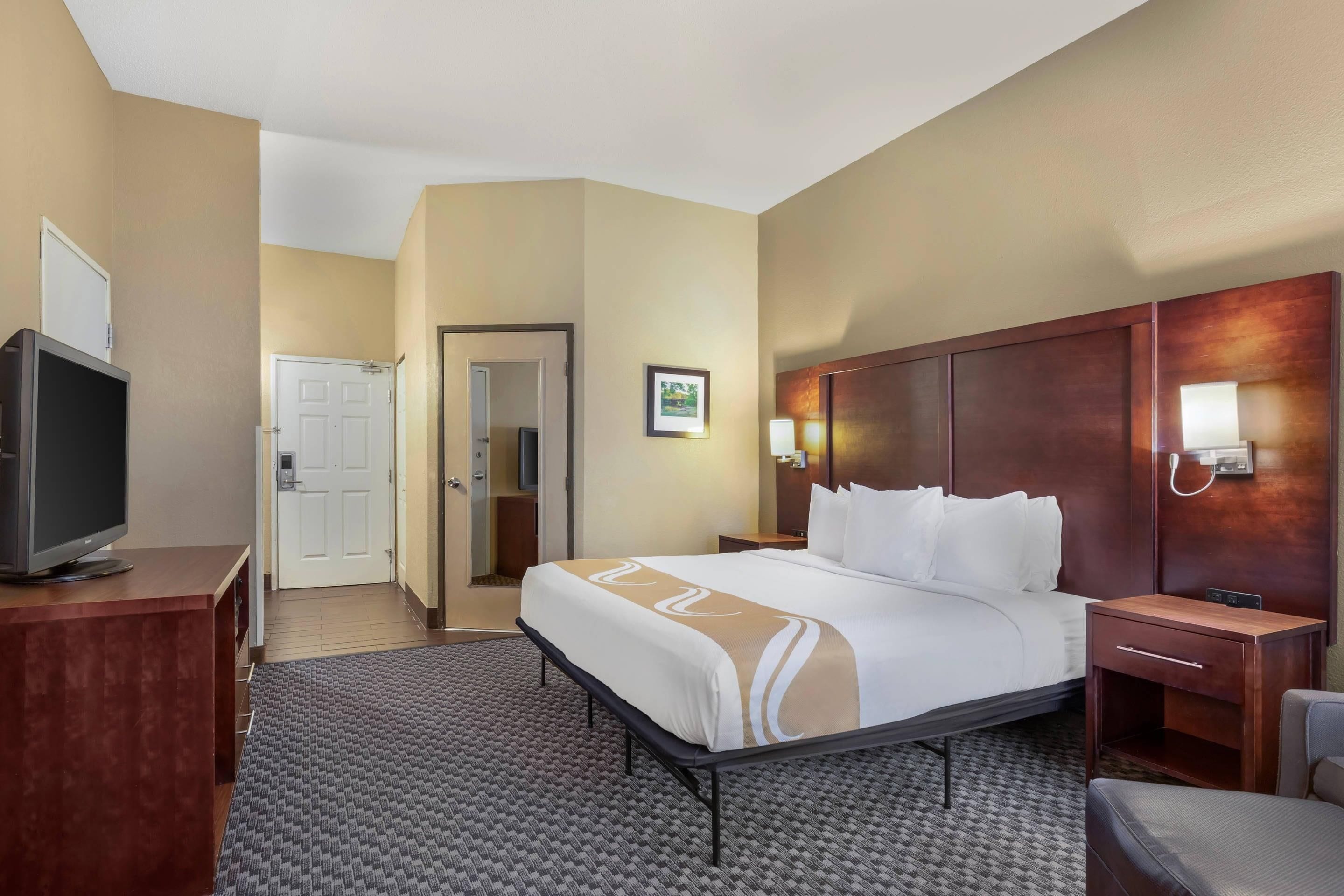 Quarto de hotel com cama, escrivaninha com televisão, cadeira, mesa de cabeceira e luminária na parede.