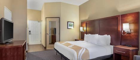 A hotel room with a bed, a desk with a television, a chair, a bedside table, and a wall-mounted lamp.