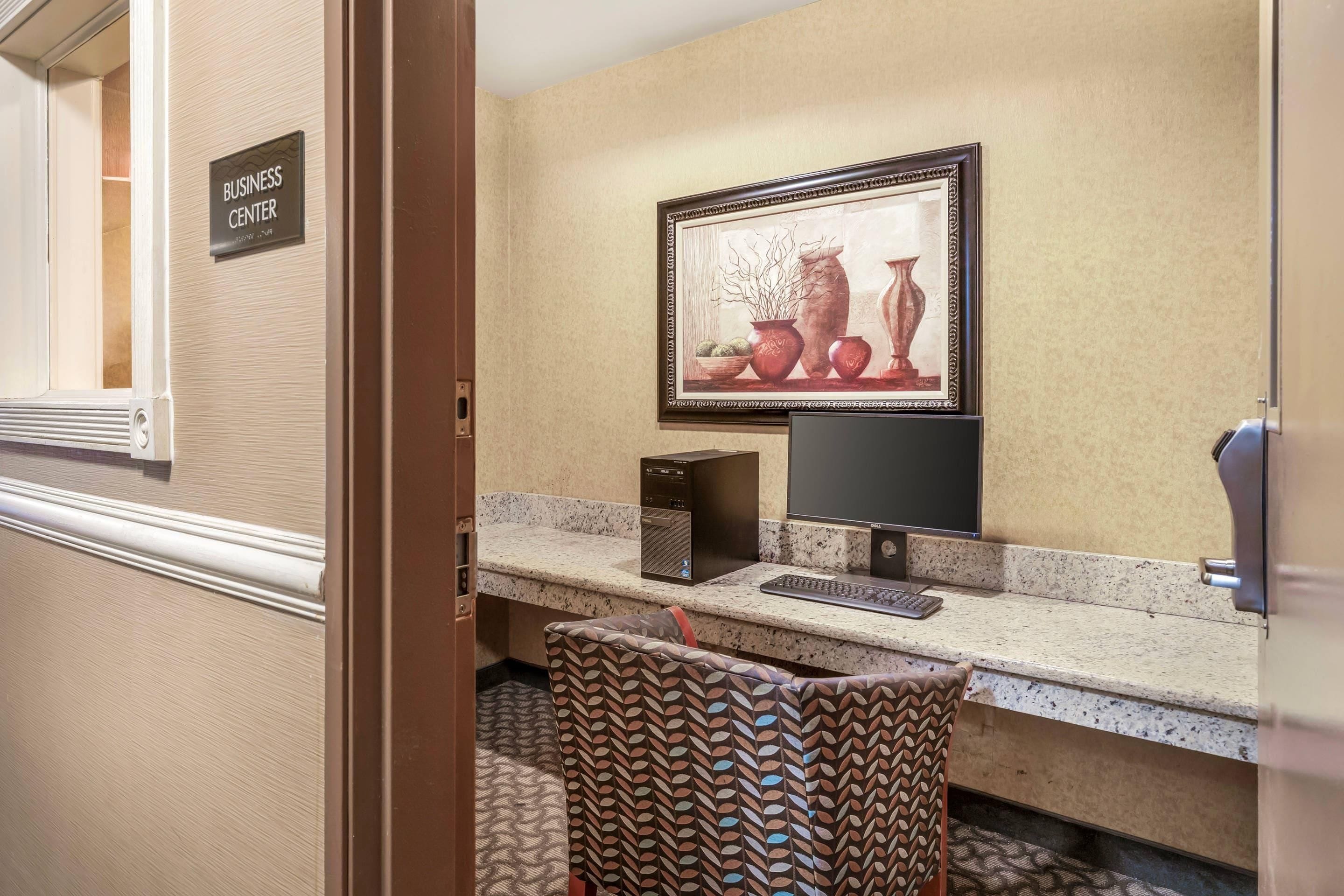 A hotel room with a business center sign, a desktop computer, a framed artwork, and a patterned chair.
