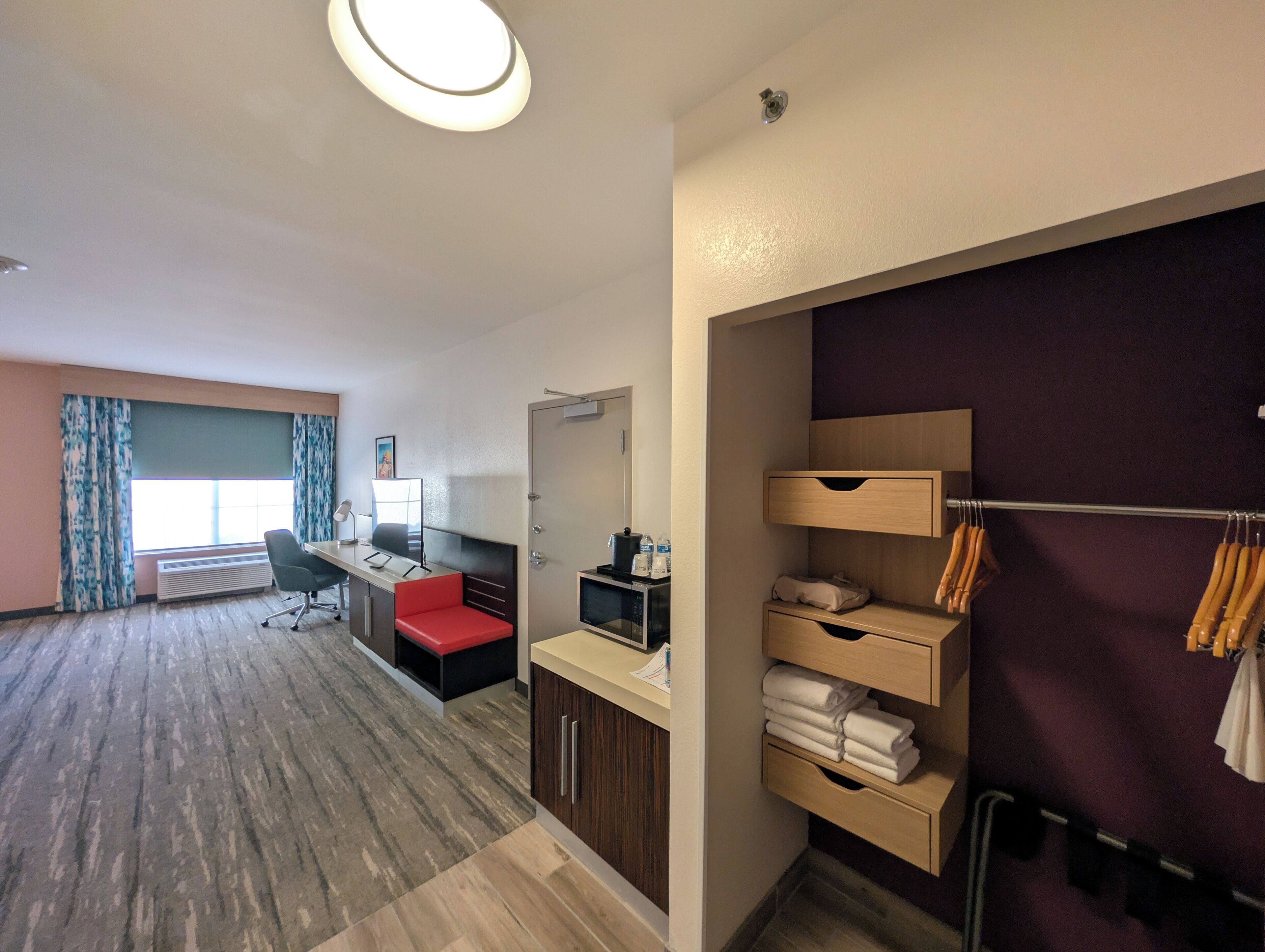 A hotel room with a desk, chair, and a bed. There is a wardrobe with hanging clothes and a shelf with towels.