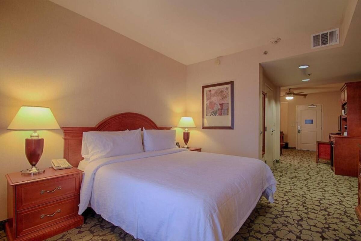 A hotel room with a bed, two bedside lamps, a nightstand, a framed picture on the wall, and a patterned carpet.
