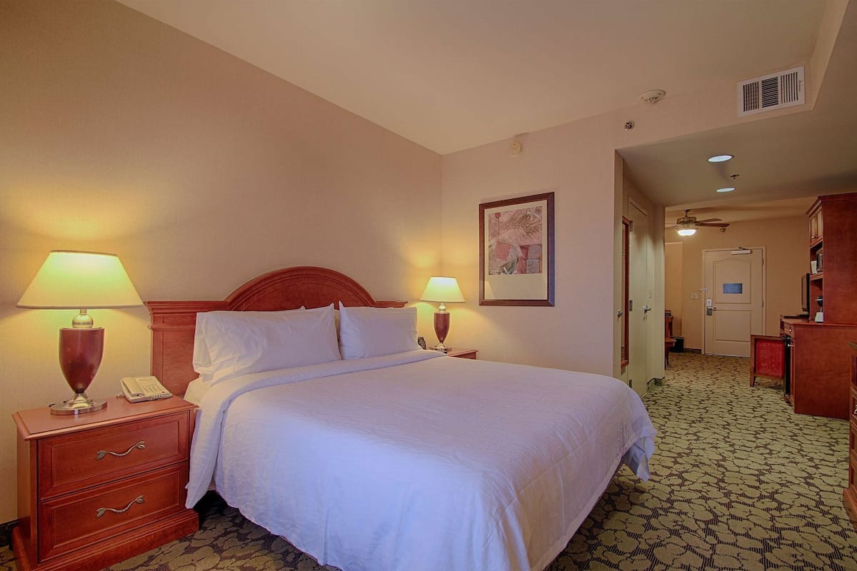 A hotel room with a bed, two bedside lamps, a telephone, a framed picture on the wall, and a door leading to another room.