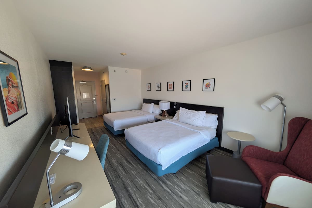 A hotel room with two beds, a desk with a lamp, a chair, a small table, and a red armchair.