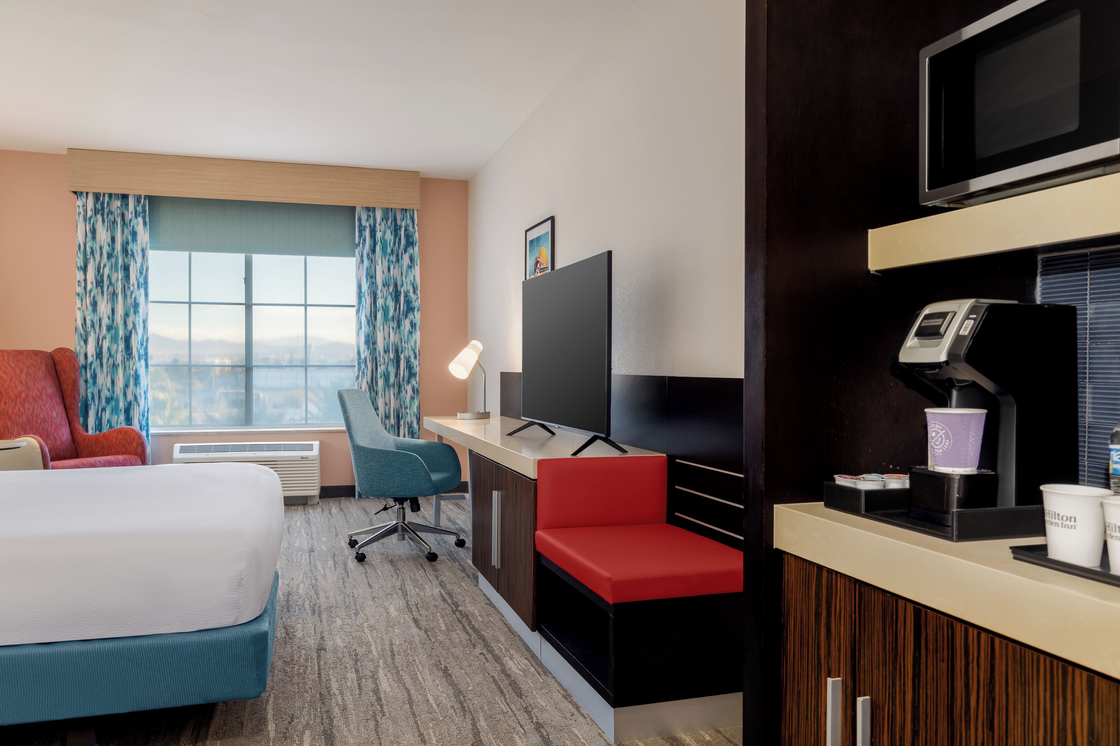 A hotel room with a bed, a desk with a computer, a TV, a coffee machine, and a window with curtains.
