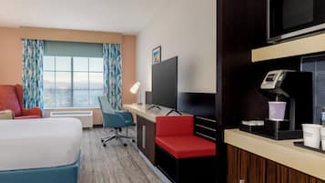 Habitación de hotel con cama, escritorio con computadora, televisor, cafetera y ventana con cortinas.