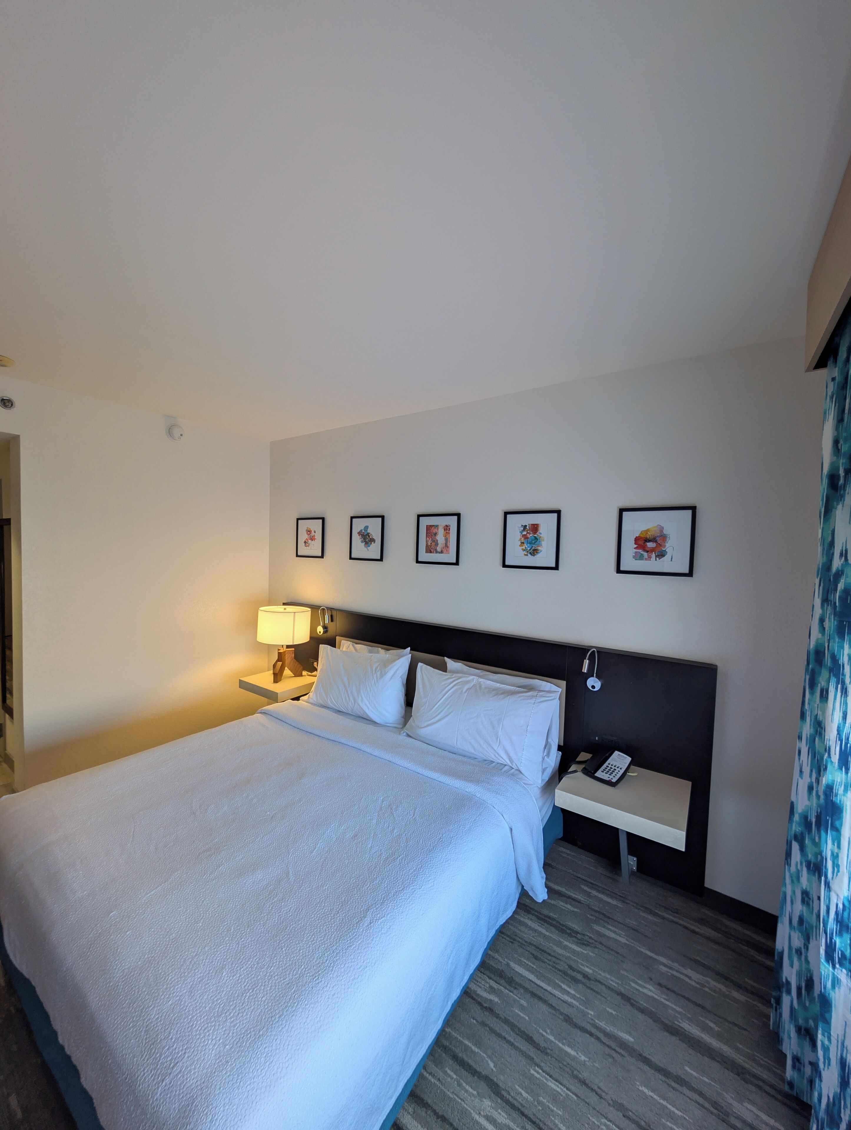 Een hotelkamer met een groot bed, een nachtkastje met een lamp, een telefoon en vier ingelijste foto's aan de muur.