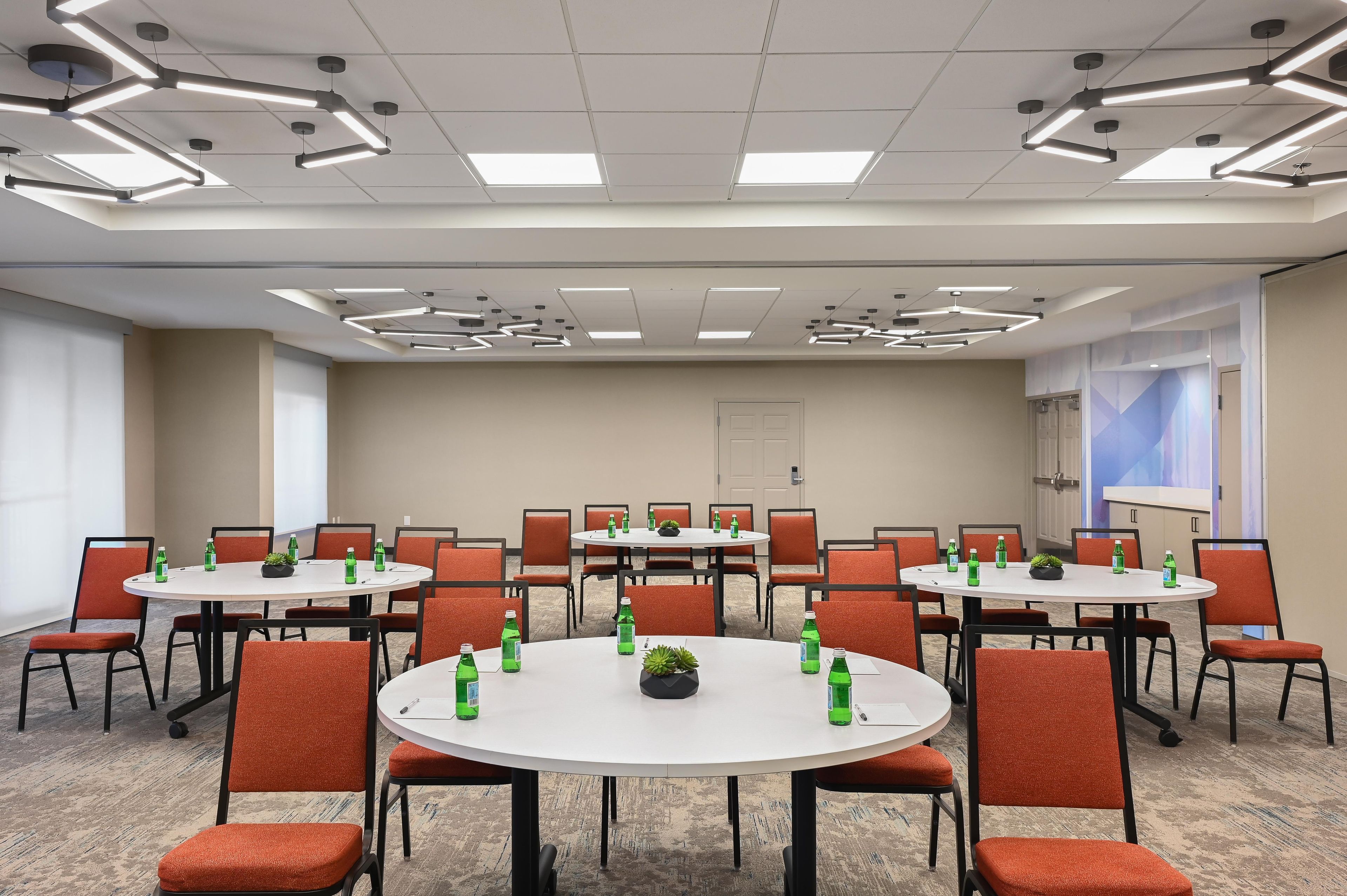 A conference room with multiple tables and chairs, equipped with ceiling lights and a projector screen.