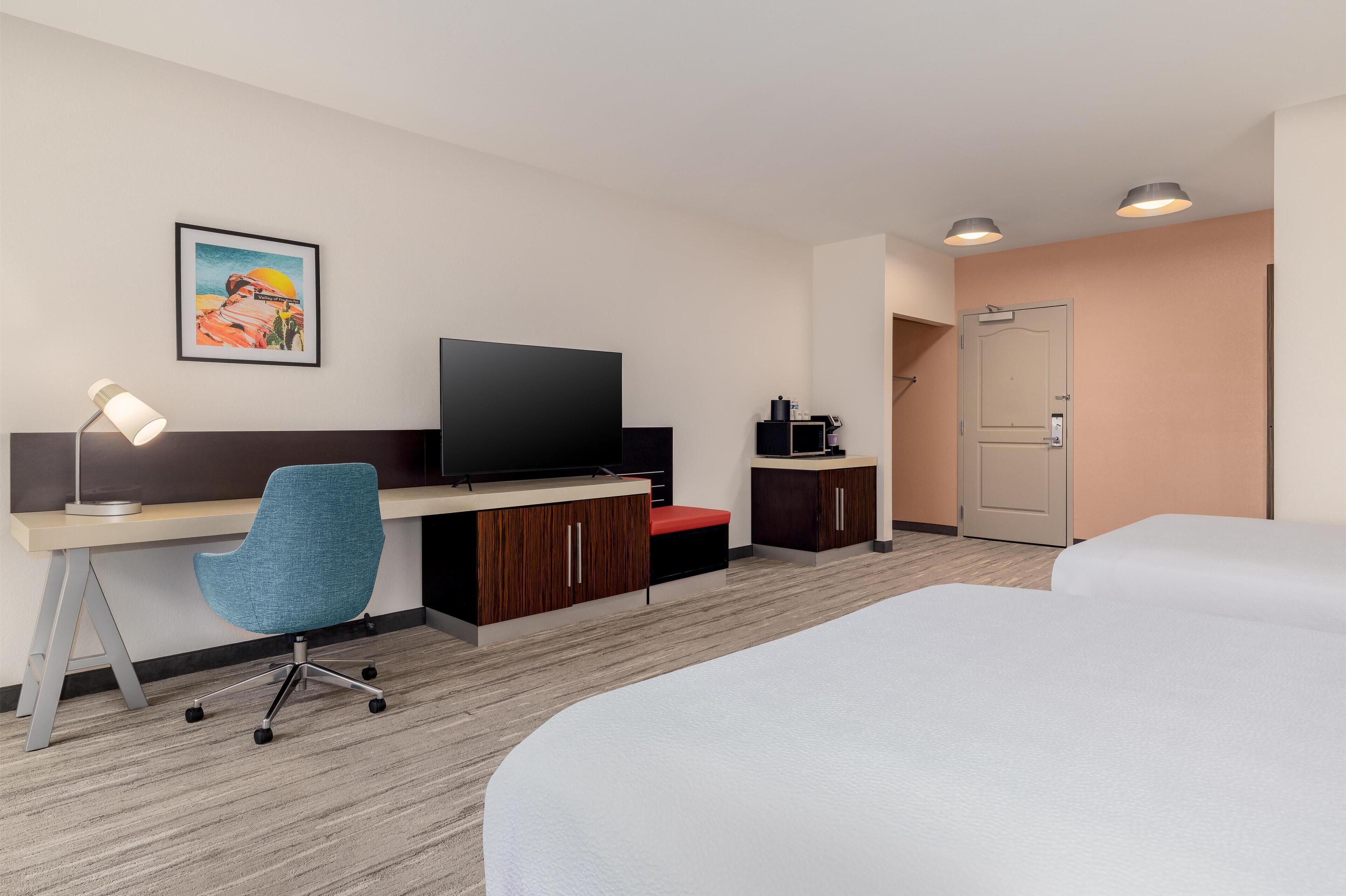 Ein Hotelzimmer mit Bett, Schreibtisch mit Stuhl, Fernseher, Kaffeemaschine und einer Tür.
