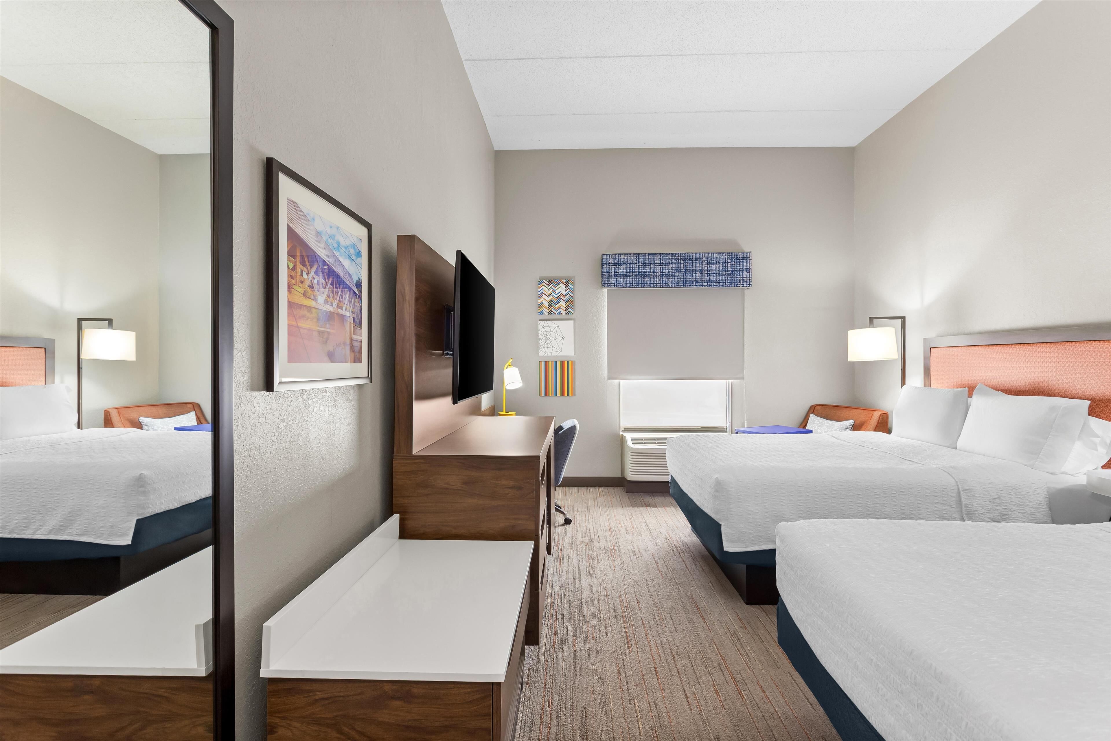 A hotel room with two beds, a desk, a chair, a TV, and a mirror.
