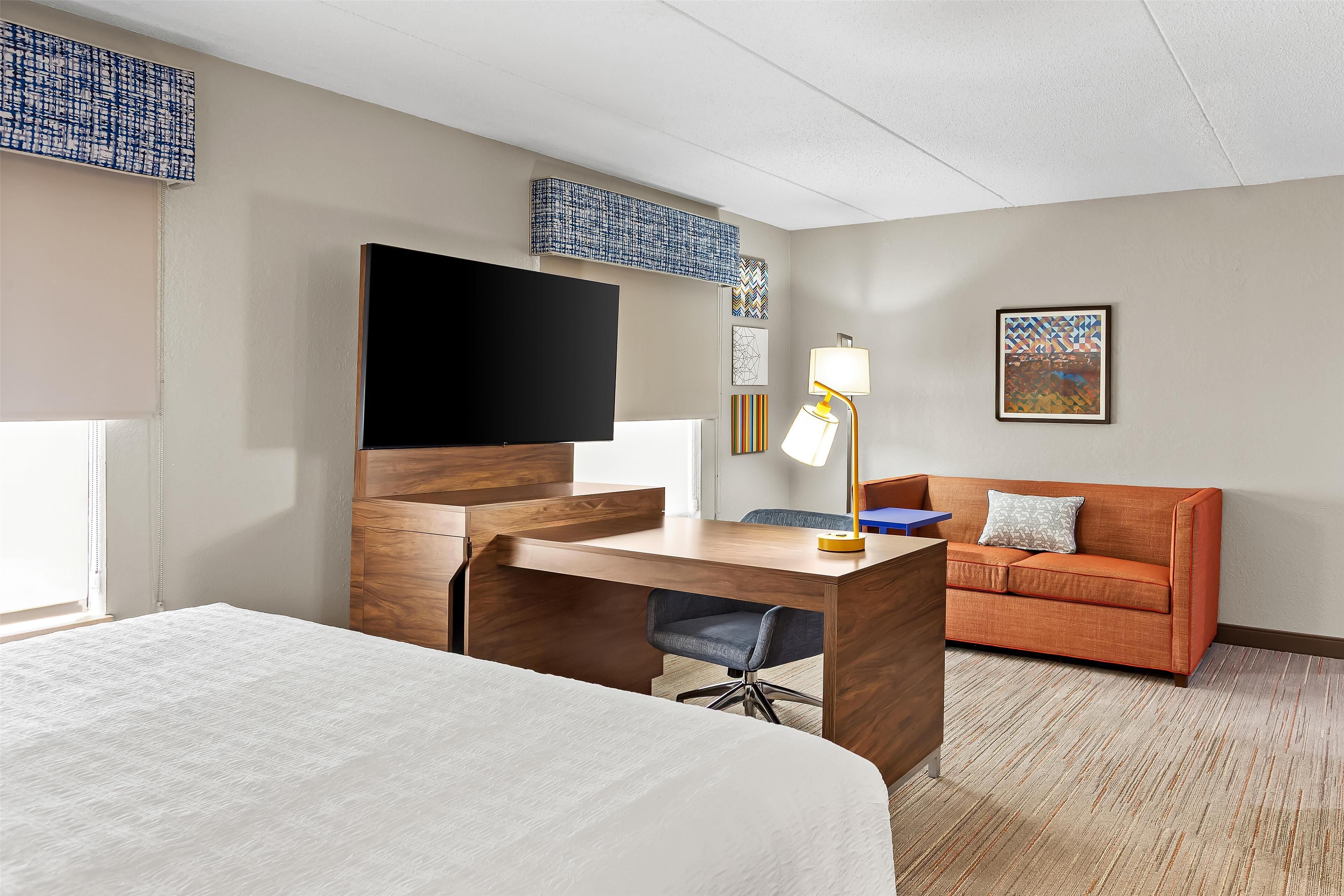 A hotel room with a bed, a desk, a sofa, a TV, and artwork on the walls.