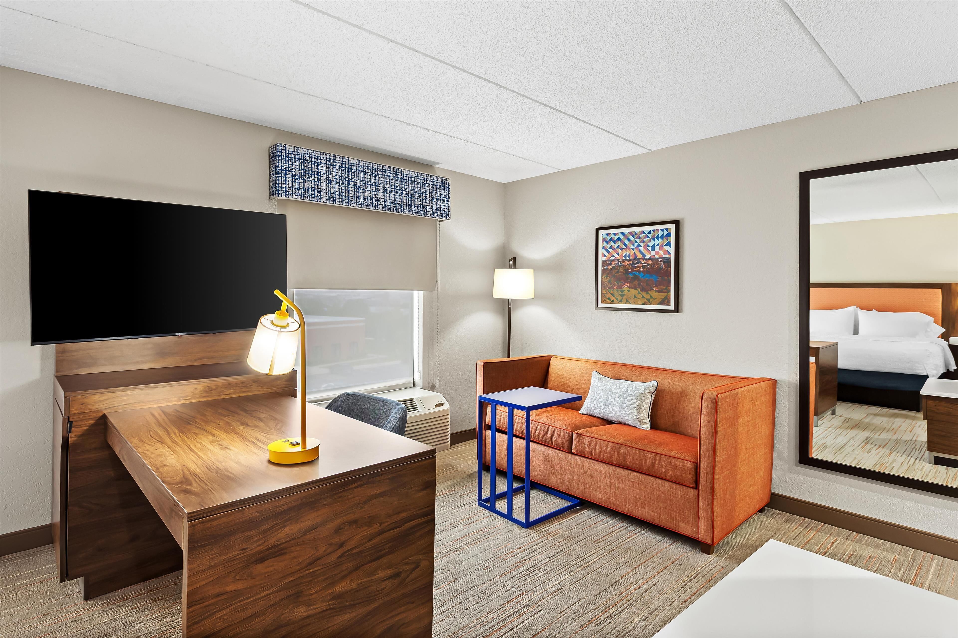 A hotel room with a wooden desk, a flat-screen TV, an orange sofa, a small blue side table, a lamp, a wall-mounted artwork, and a large mirror reflecting the bed.