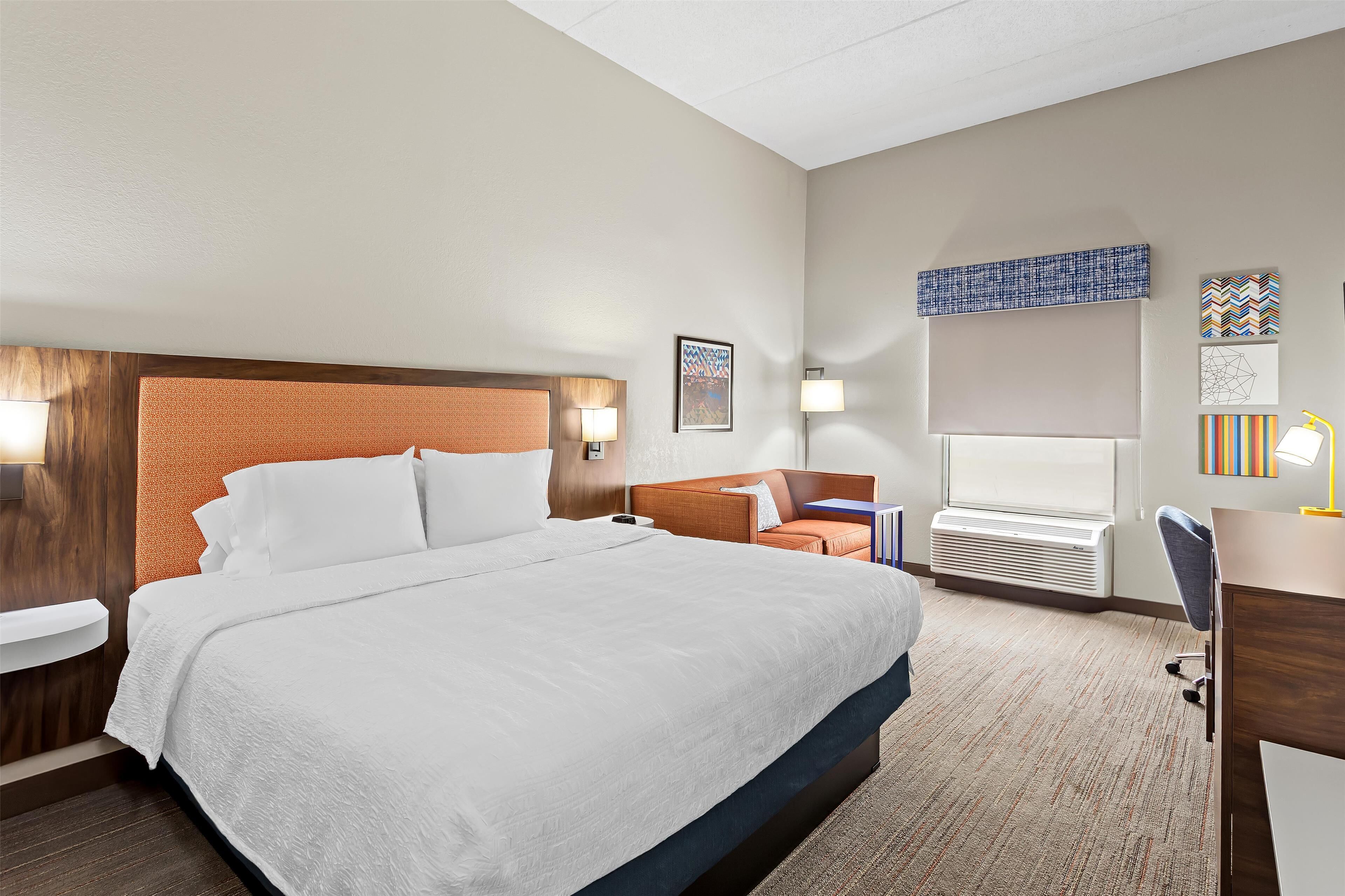 A hotel room with a large bed, a desk, a chair, a small table, and a wall-mounted air conditioner.