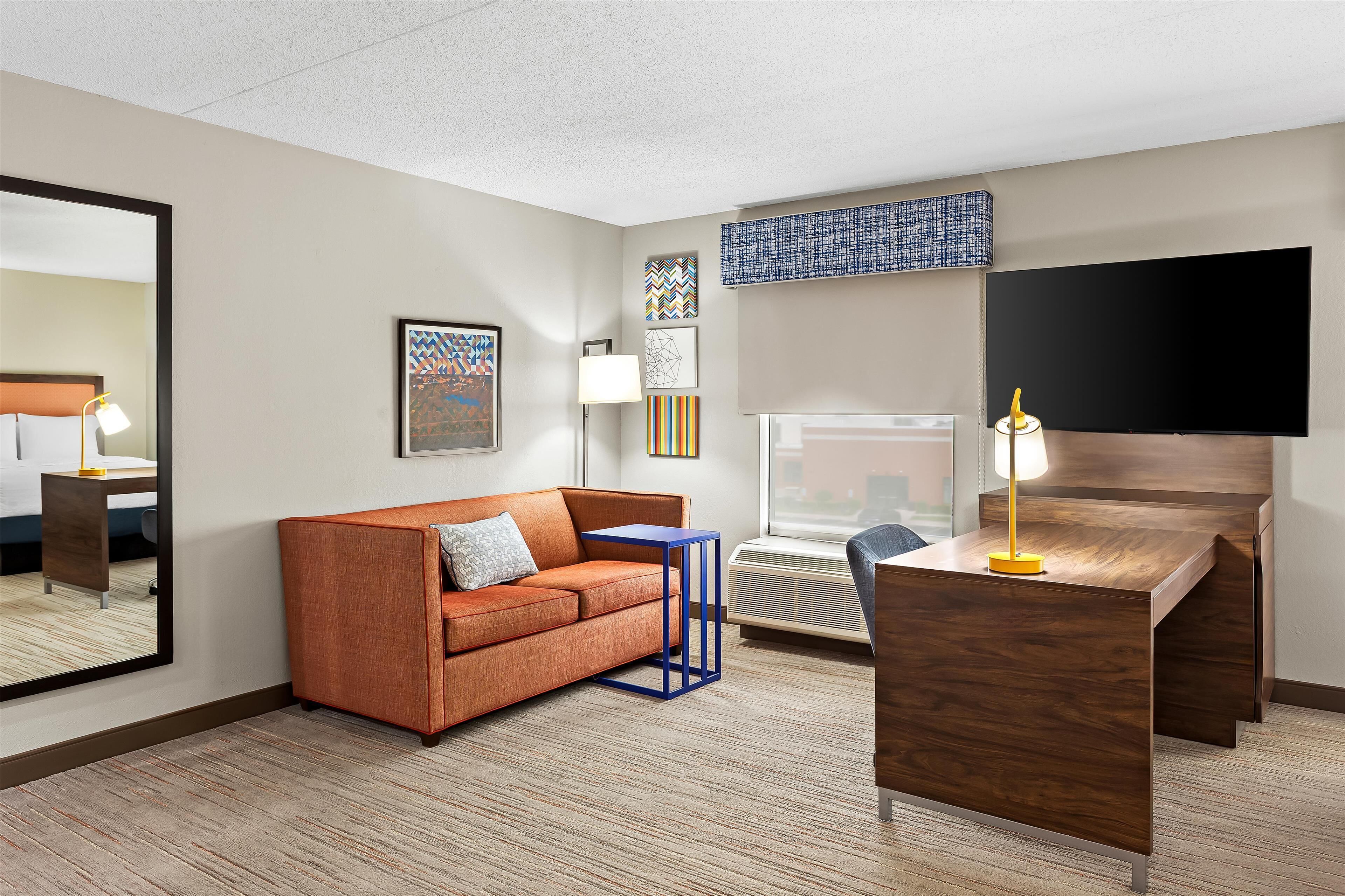 Una habitación de hotel con un escritorio de madera, sofá naranja y un espejo grande.