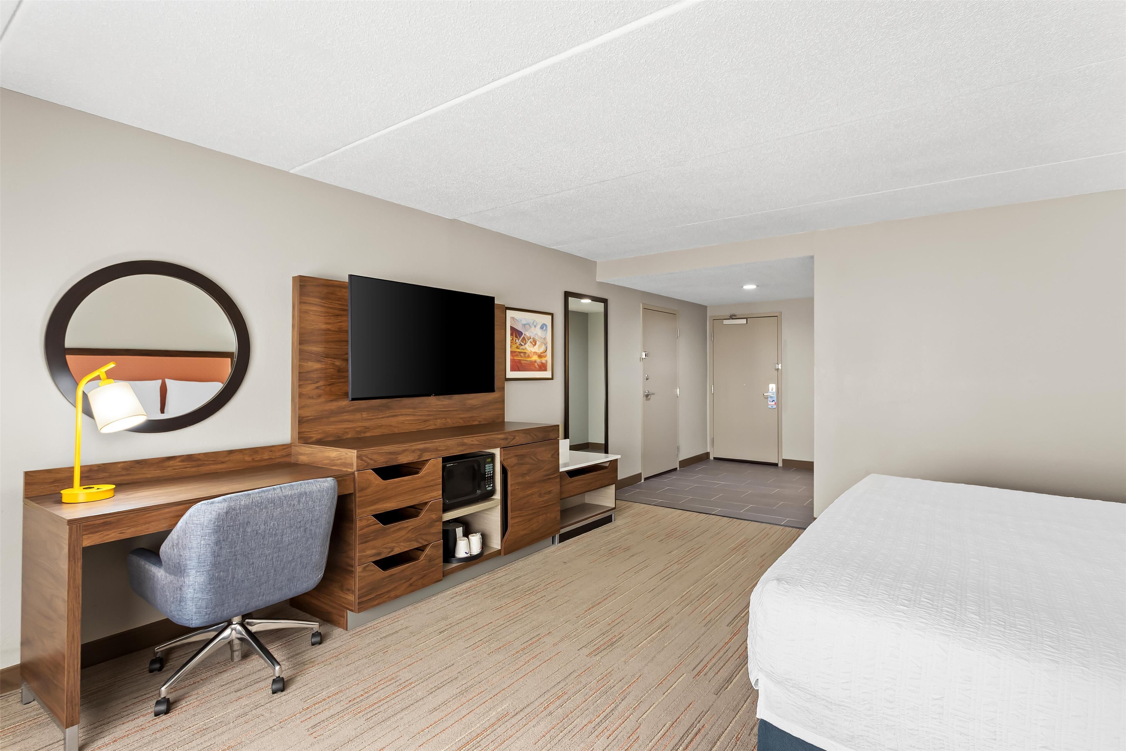 A hotel room with a wooden desk, a flat-screen TV, a large mirror, a bed, and a door leading to another room.
