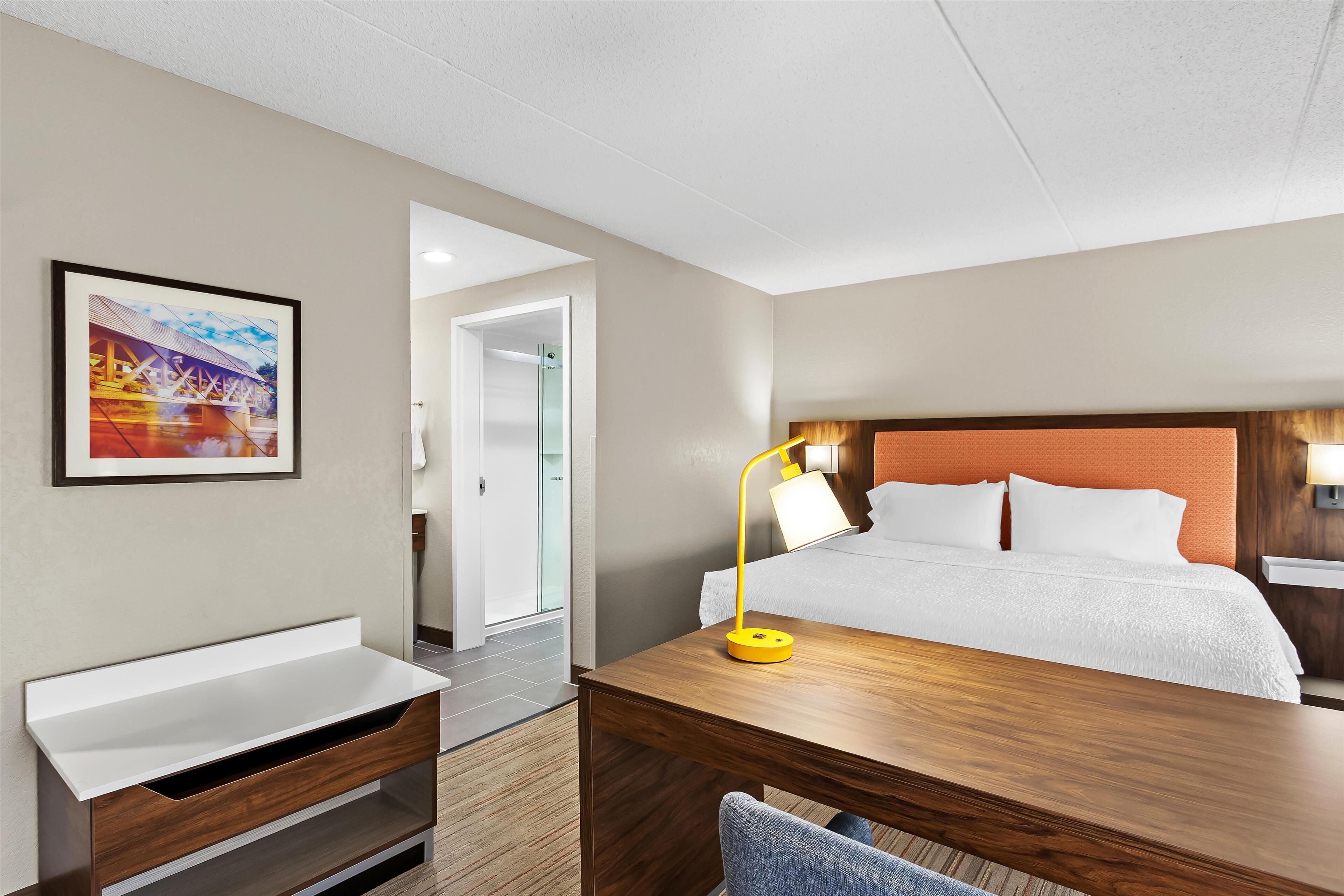 Habitación de hotel con cama, escritorio, silla, mesita de noche, lámpara y un cuadro en la pared.