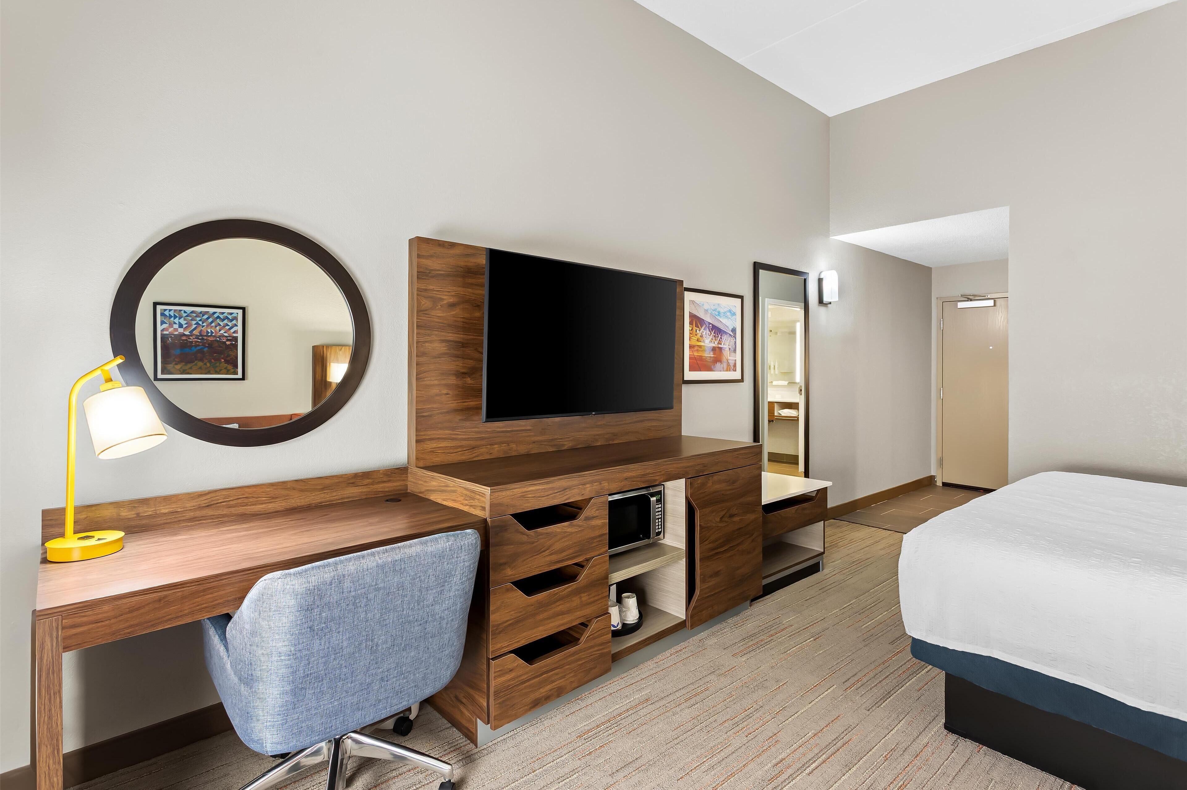 Ein Hotelzimmer mit Holzschreibtisch, einem Sessel, einem Bett, einem Fernseher und einem Spiegel.