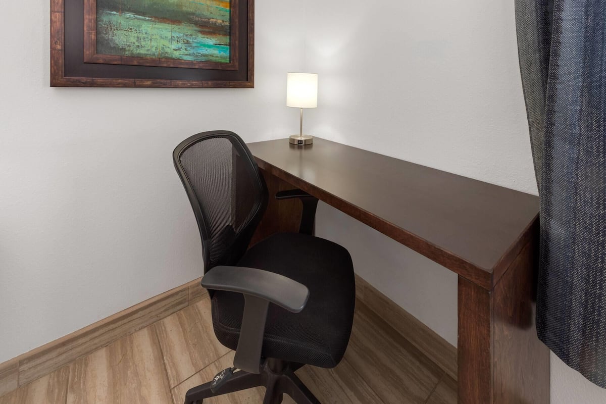 A corner office with a desk, chair, lamp, and a framed painting on the wall.