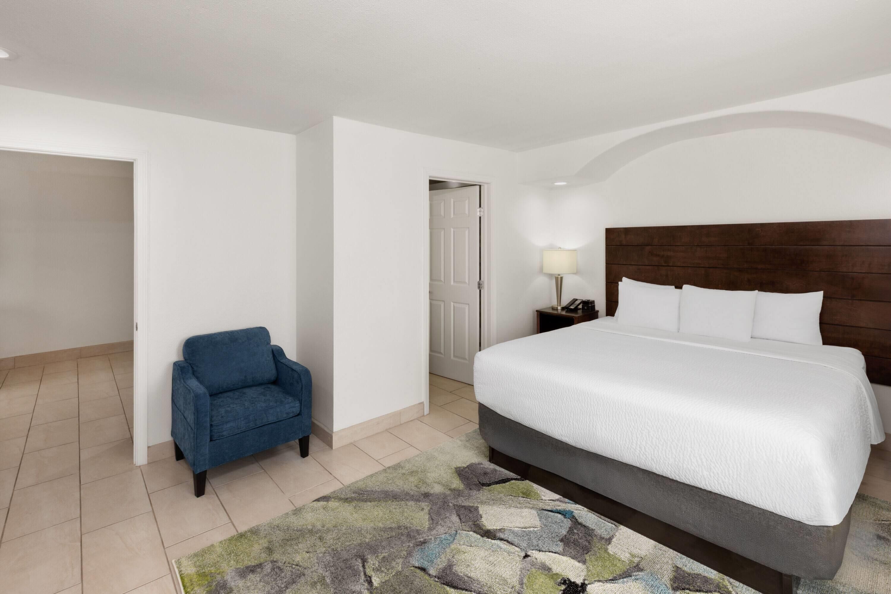 A hotel room with a bed, a blue chair, a nightstand with a lamp, and a door leading to another room.