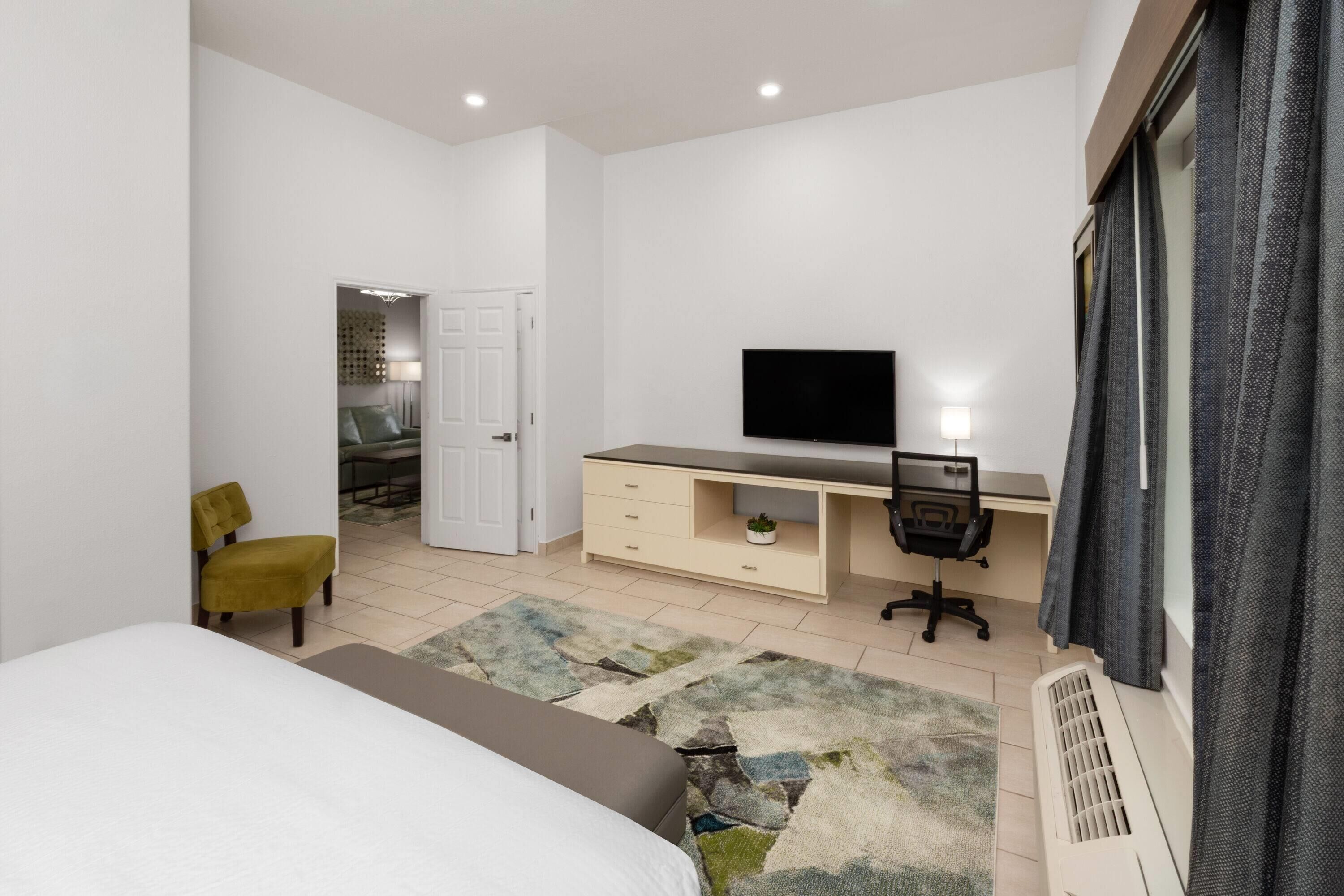 Un dormitorio moderno con cama, escritorio, silla, televisor y una puerta que da al baño.