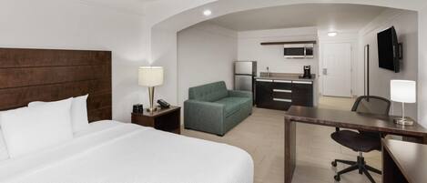 Quarto de hotel com cama grande, escrivaninha, poltrona verde, cozinha compacta e televisão na parede.