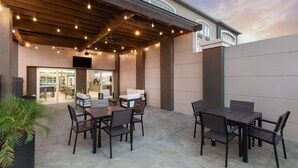 An outdoor patio with tables and chairs, a TV mounted on the wall, and a view into the building. - La Quinta Inn & Suites by Wyndham Laredo Airport (Laredo)